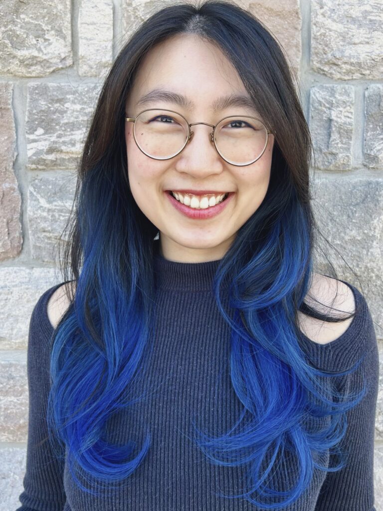 Photo of Flora Pan against a brick wall backdrop. She has long black and blue hair with round glasses in the photo, and is wearing a dark grey sweater.